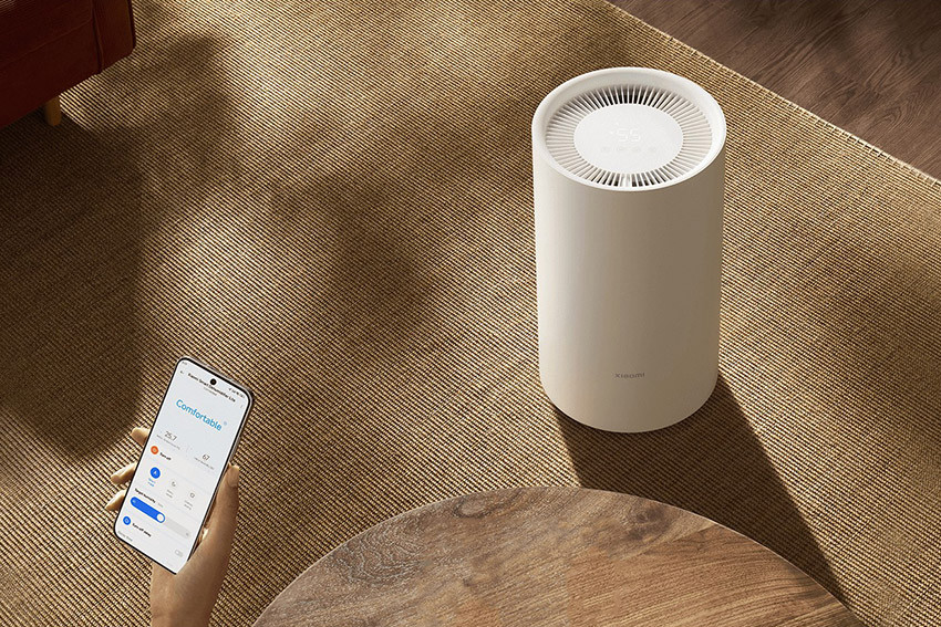Xiaomi Smart Dehumidifier Lite na dywanie obok stolika i telefon w dłoni z aplikacją Xiaomi Home